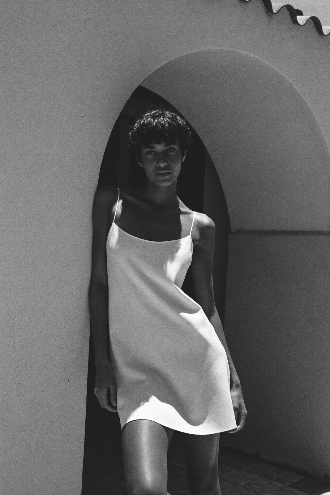 Monochrome standing portrait in a slip dress under a limewash archway, hard sun