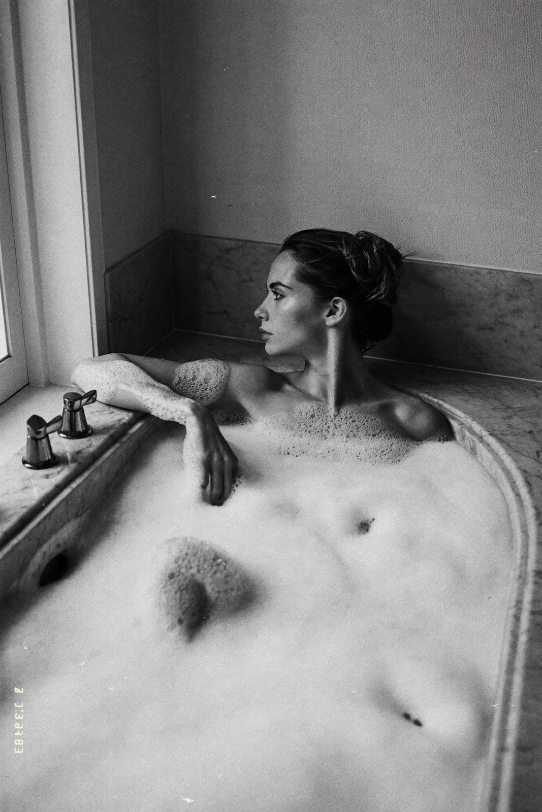 Monochrome portrait in a deep marble bath, foam to the collarbones, window light