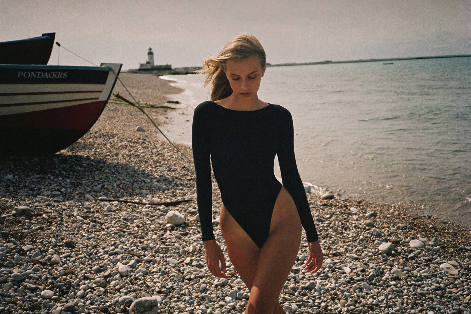 Walking barefoot on Dungeness shingle in a black swimsuit, fishing boat and lighthouse behind