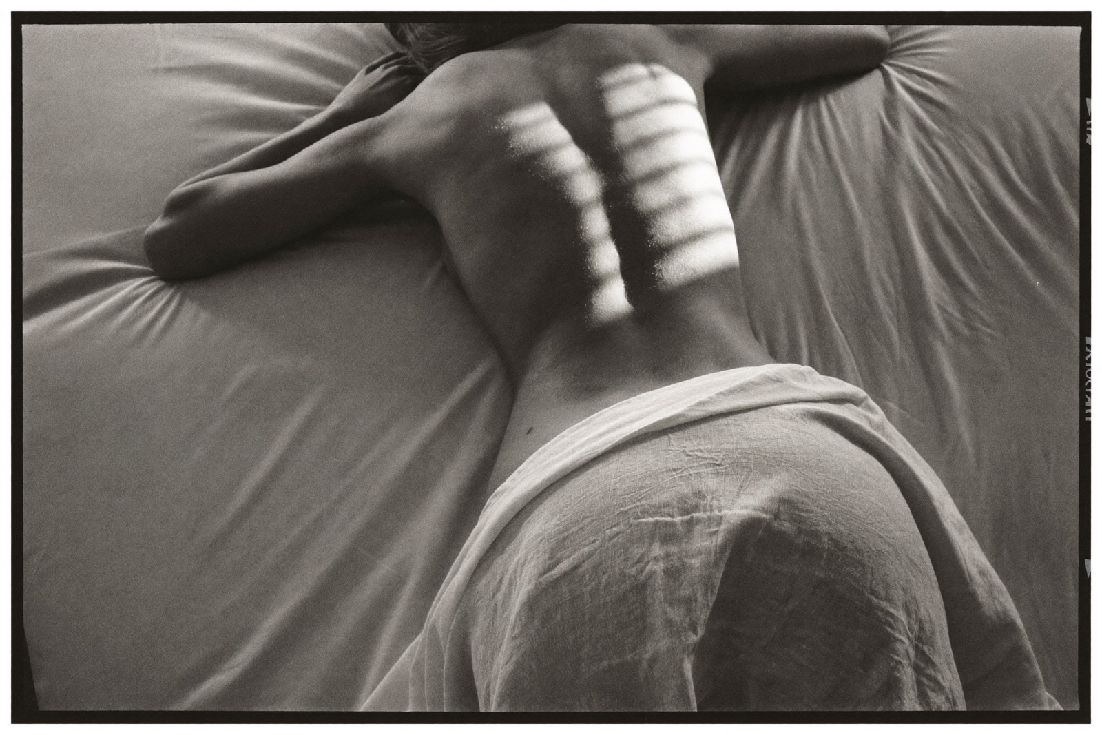 Monochrome, lying face down on white linen with a sheet across the hips, raking window light