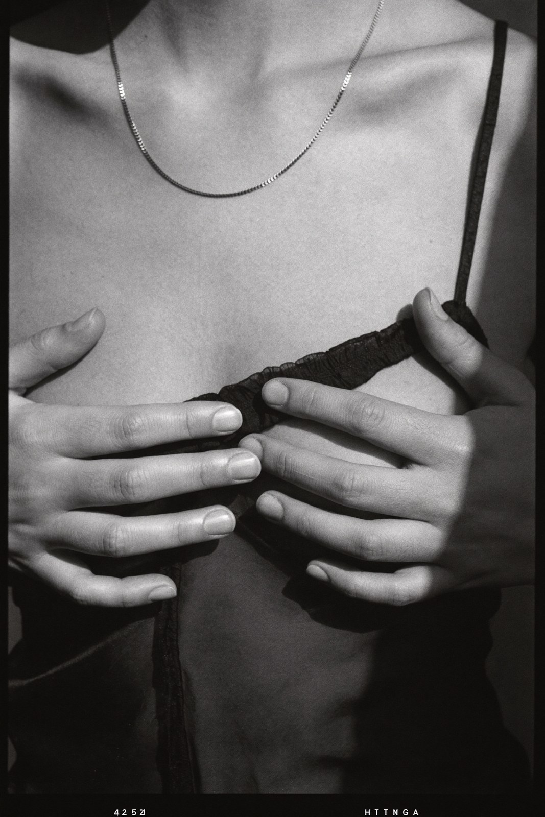 Monochrome close crop of a collarbone, hands and a thin gold chain, blind shadows
